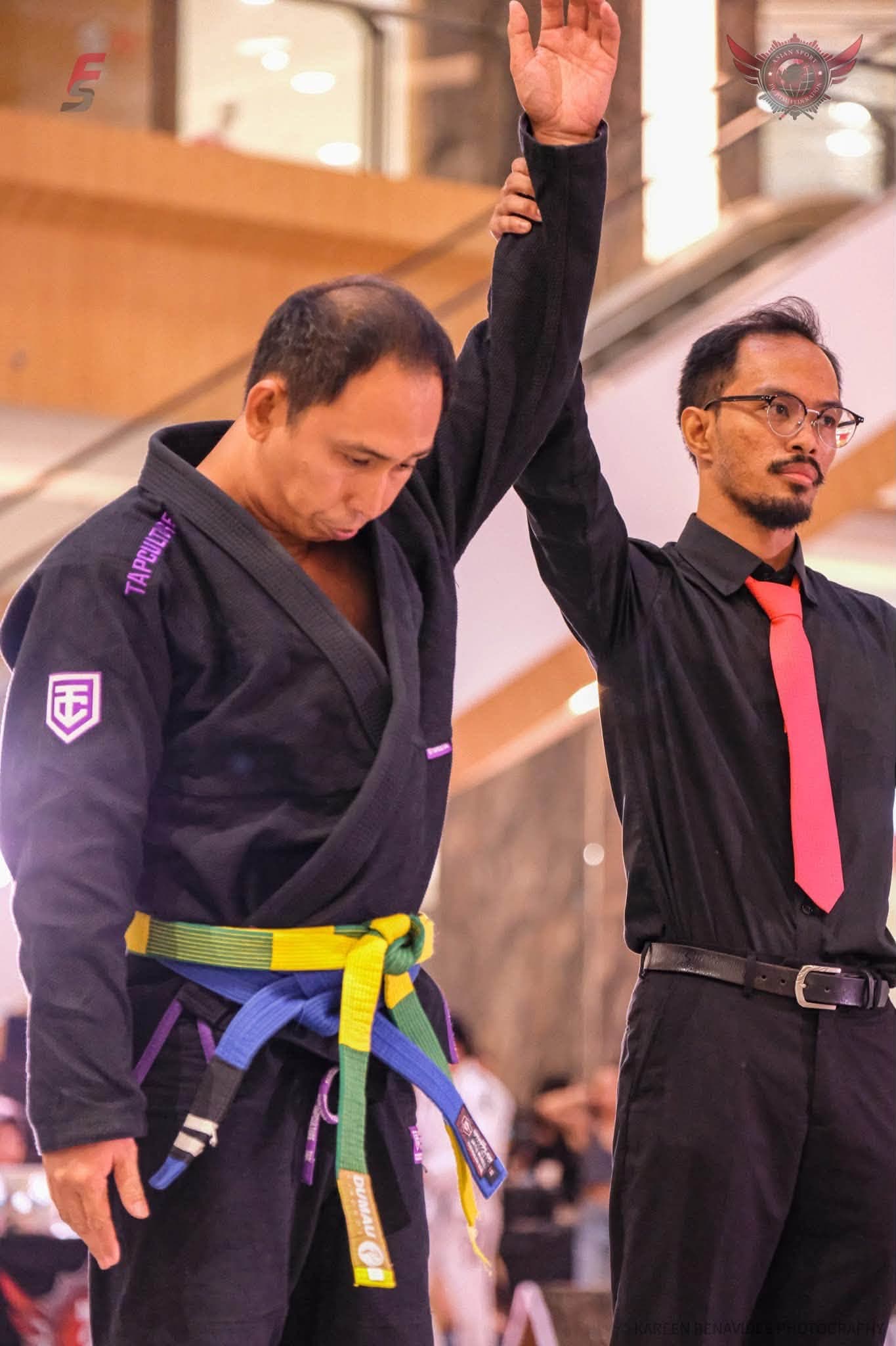 A Kabkad athlete in a black gi has his hand raised by the referee after winning a match